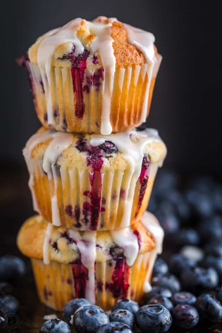 These lemon blueberry muffins are bursting with fresh blueberry flavor. This blueberry muffin recipe has a soft and moist crumb and they puff up perfectly. The easy lemon glaze recipe makes them completely irresistible and they always disappear fast! | natashaskitchen.com Blueberry Muffins glazed and stacked surrounded by fresh blueberries
