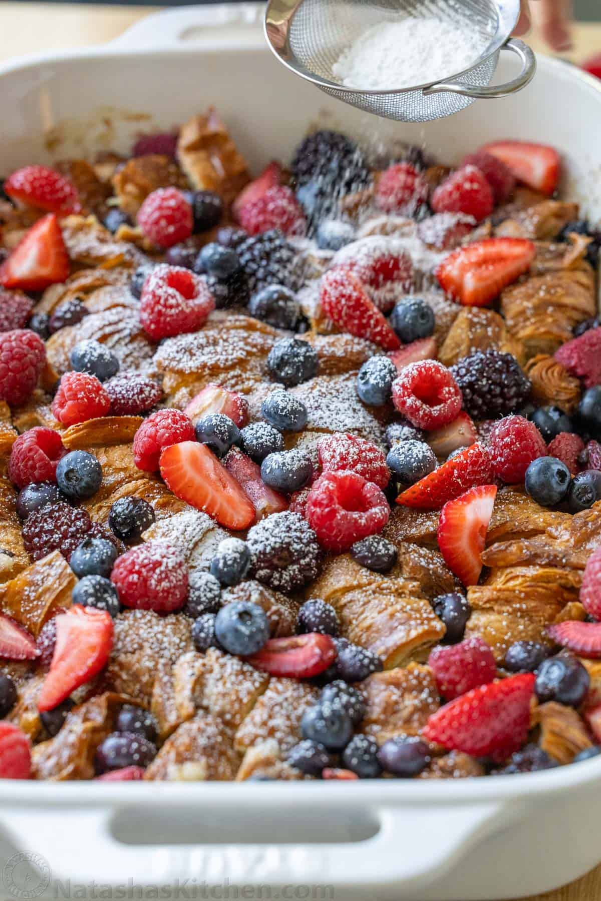 Easy Croissant French Toast Casserole with fresh berries is the best crowd-pleasing breakfast recipe. Refrigerate overnight for easy serving. Dusting powdered sugar over croissant French Toast Casserole in a white dish