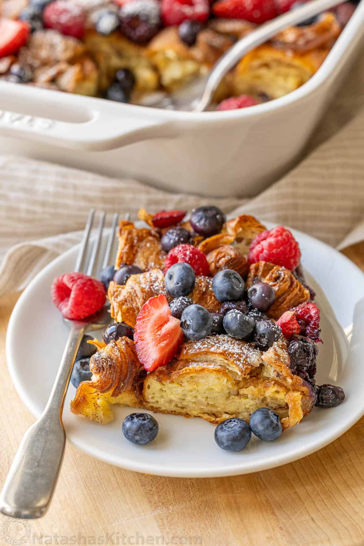 Easy Croissant French Toast Casserole with fresh berries is the best crowd-pleasing brunch. Refrigerate overnight for a make-ahead breakfast. Portion of Croissant French Toast Bake on a white plate with berries on top and a fork
