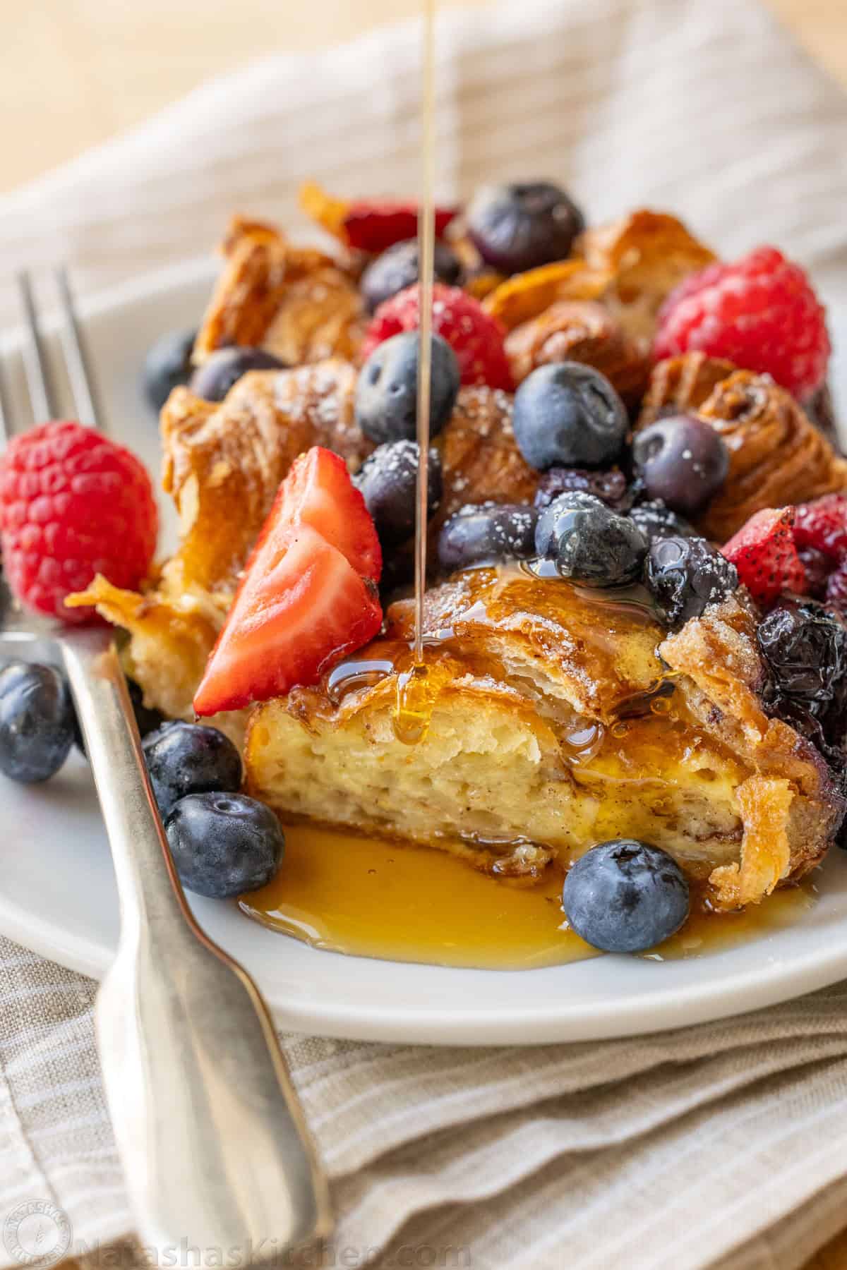 Easy Croissant French Toast Casserole with fresh berries is the best crowd-pleasing breakfast recipe. Refrigerate overnight for easy serving. syrup drizzled on Croissant French Toast Bake with fresh berries on top and a fork to eat