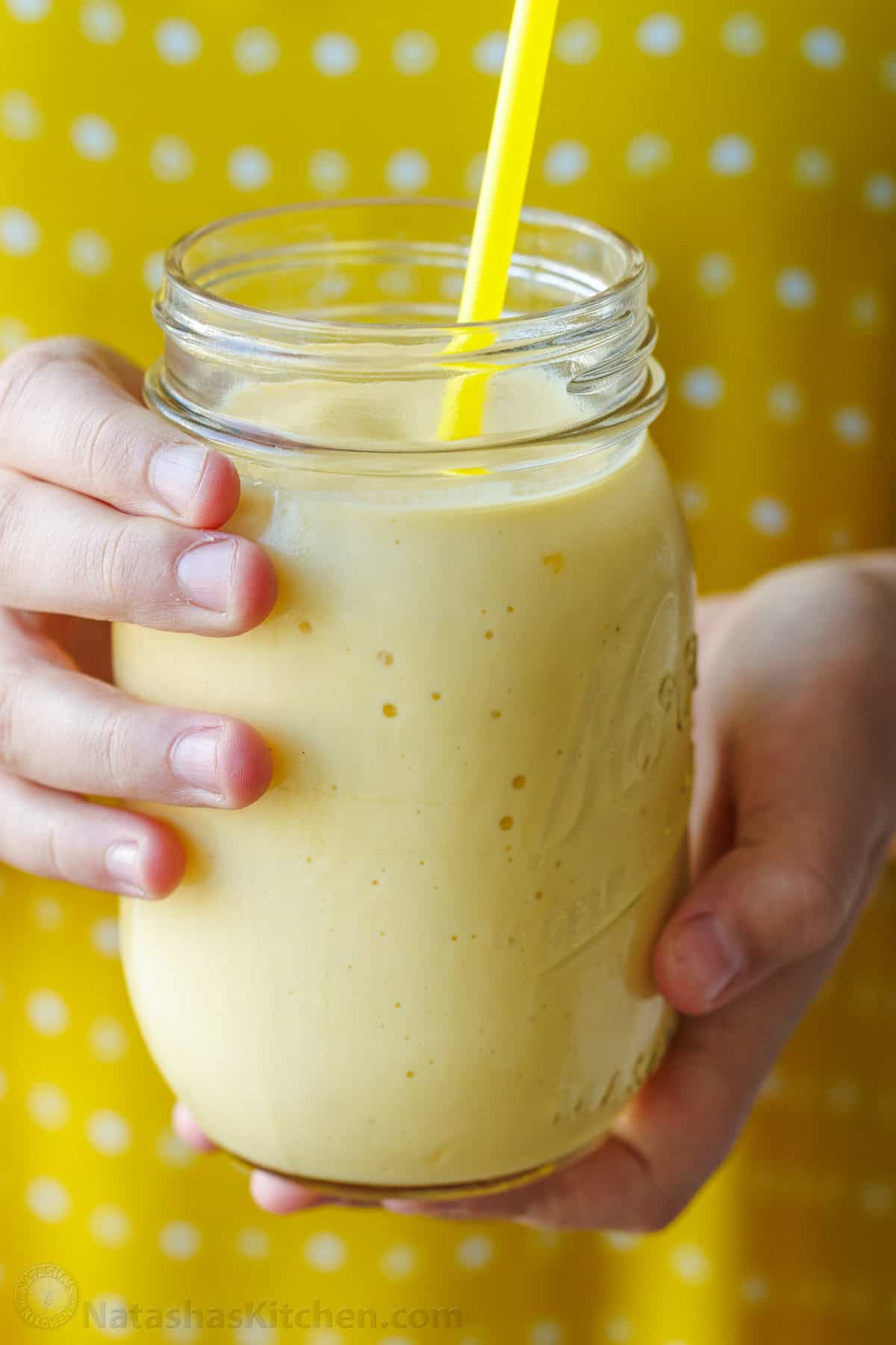 Easy Tropical Mango Smoothie recipe uses just 6 ingredients, like mango, lime, banana, and Greek yogurt, and takes under 5 minutes to make! hands holding a yellow mango smoothie in a mason jar with yellow polka-dot background
