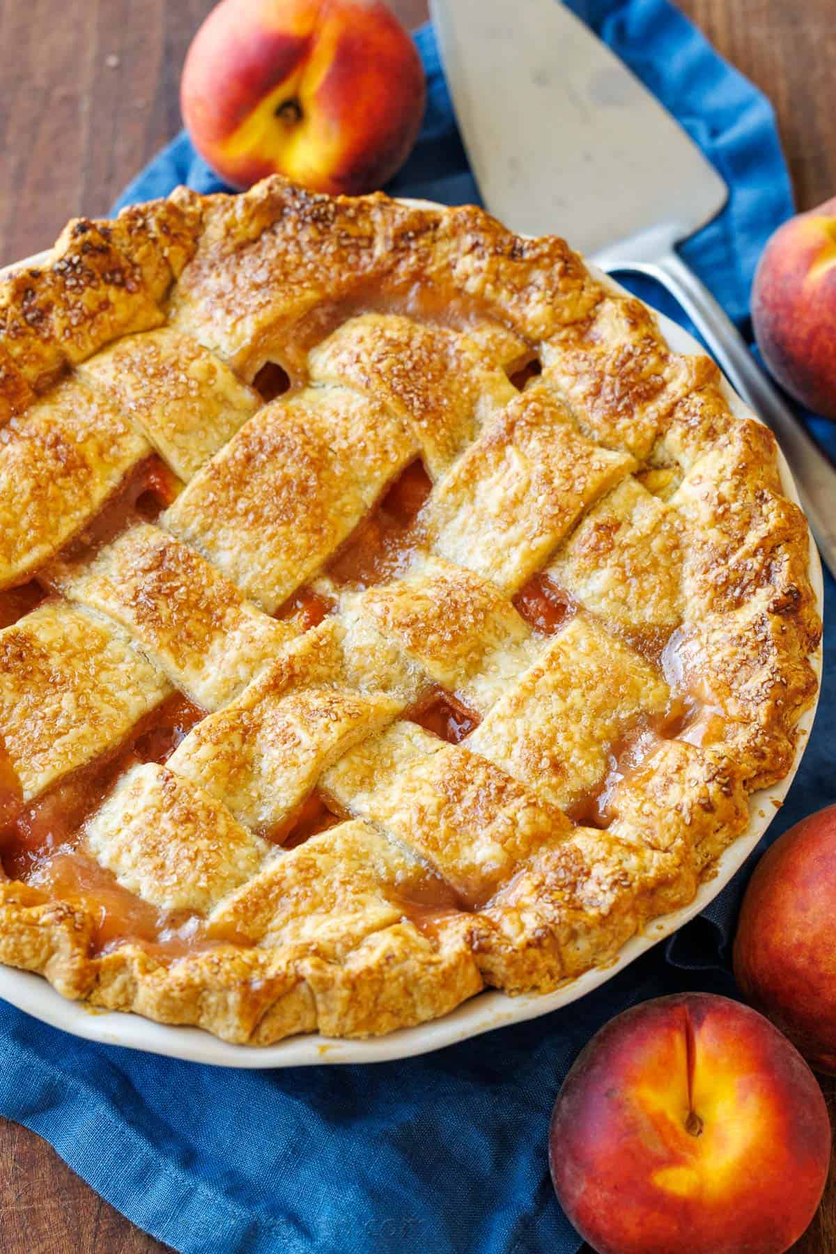 Easy homemade Peach Pie Recipe with pie crust made from scratch. This fool-proof recipe tastes amazing with ripe peaches. Gets rave reviews! Peaches surrounding a homemade peach pie with browned crust and pie server
