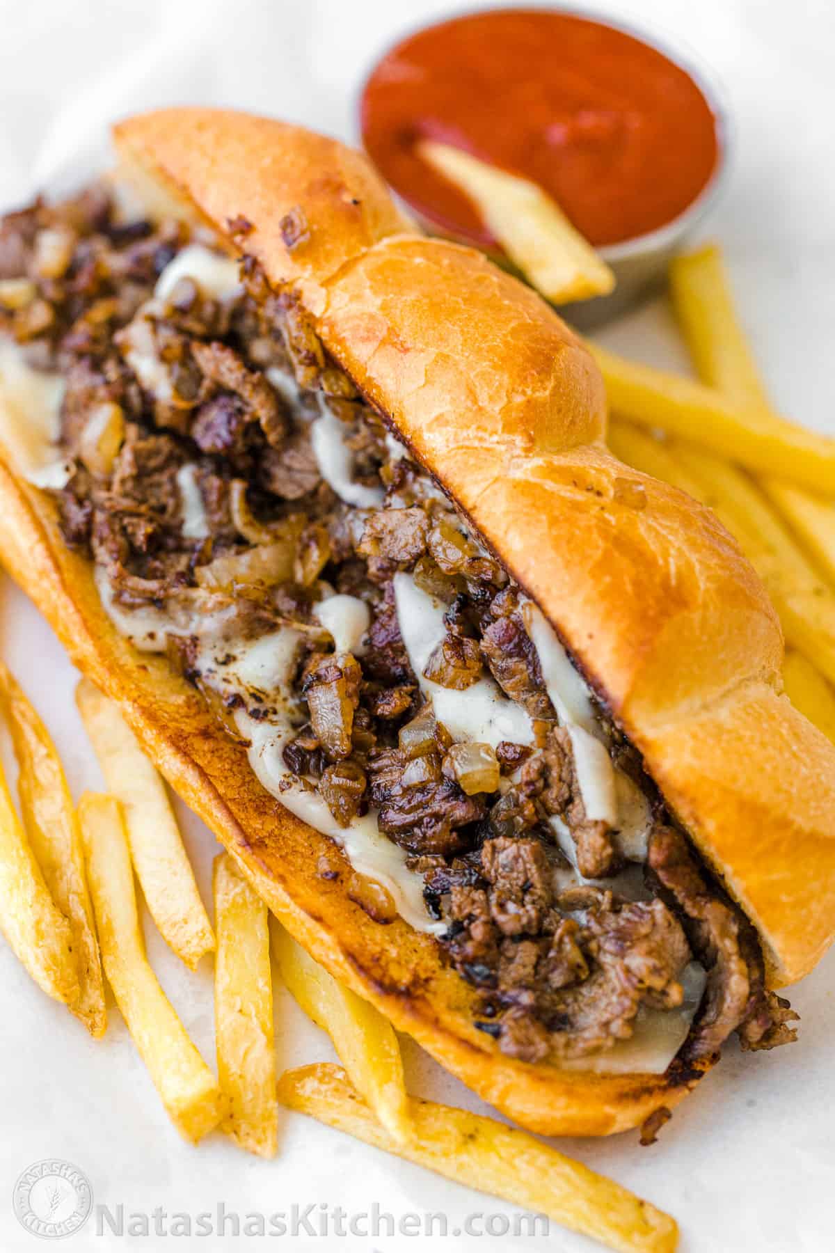 Philly Cheesesteak with tender ribeye steak, melted provolone, and caramelized onion in a garlic butter roll. Easy Philly Cheesesteak Sandwich video how-to. #phillycheesesteak #phillycheesesteakrecipe #phillycheesesteaksandwich #natashaskitchen #cheesesteak #sandwich Philly cheesesteak sandwich served in a bun with fries and ketchup
