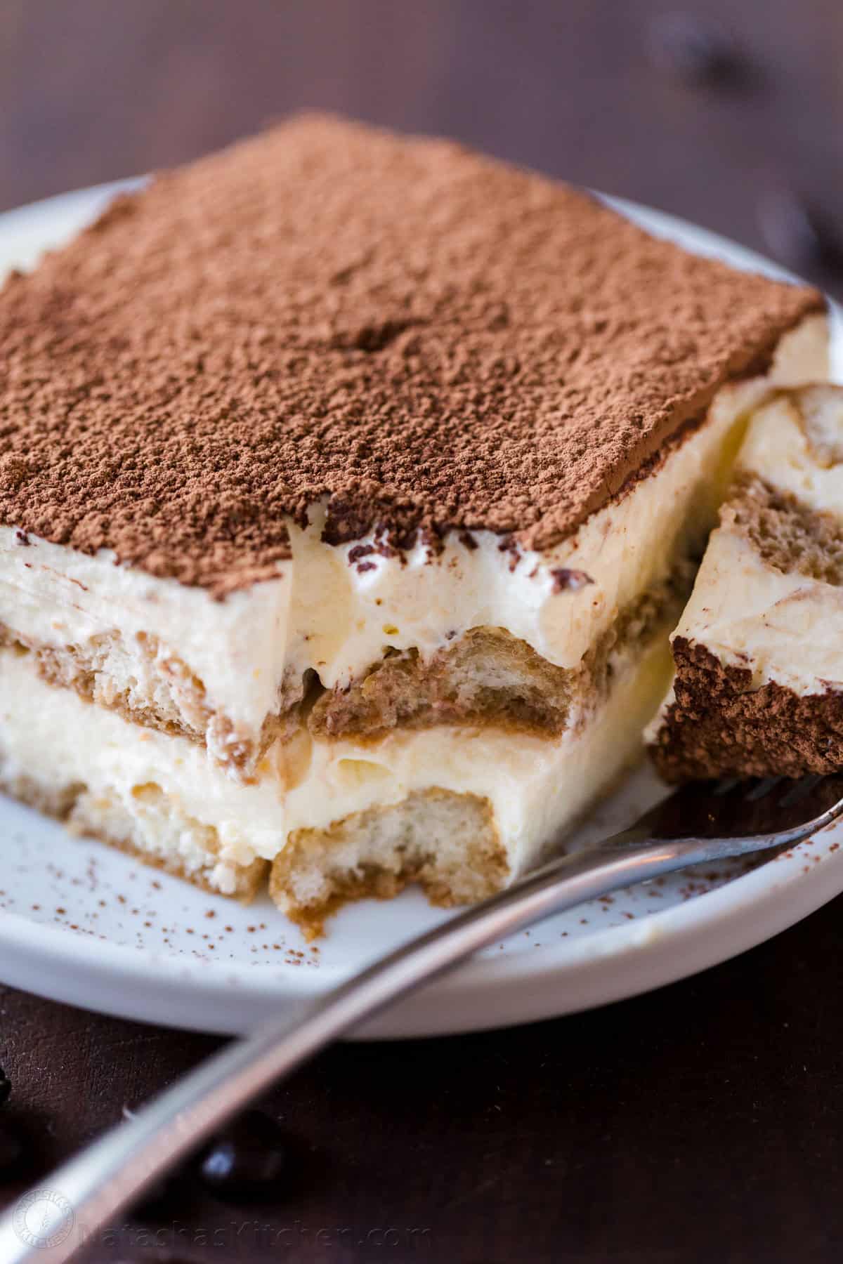 The best Classic Italian Tiramisu recipe with layers of coffee-soaked ladyfingers & decadent cream is so easy! Try my no-bake dessert recipe! a slice of homemade tiramisu cake on a plate with a bite taken out of the corner with a fork